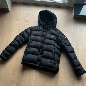 Svart dunjacka från Peak Performance - Säljer en svart dunjacka från Peak Performance med huva och dragkedja. Jackan har puffad design, snörning i nederkant och långa ärmar. Perfekt för kalla dagar och har en clean look med diskret logga på bröstet och ärmen. Andvänd 1 gång som ny. Storlek S.