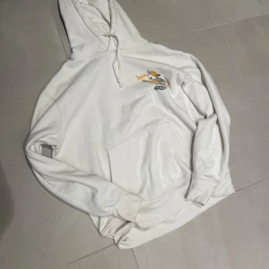 Vit hoodie Aranciata med tryck - Vit hoodie från Aranciata med stor orange och svart tryck på ryggen och mindre tryck på bröstet. Tröjan har huva med snörning, magficka och långa ärmar. Materialet känns mjukt och är troligen bomull. Perfekt för dig som gillar coola grafiska prints.