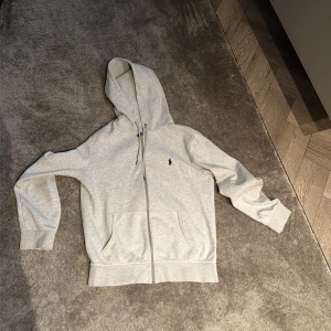 Ljusgrå hoodie från Polo Ralph Lauren - Säljer en ljusgrå hoodie från Polo Ralph Lauren i storlek S. Tröjan har huva med snörning, dragkedja framtill och två fickor. Klassisk logga broderad på bröstet. Perfekt för chill dagar och enkel att matcha med jeans eller joggers.