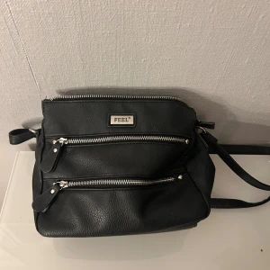 Svart axelväska från FEEL med dragkedjor - Svart axelväska i syntetmaterial från FEEL med flera fack och silverfärgade dragkedjor. Väskan har justerbar axelrem och snyggt mönstrat innerfoder. 