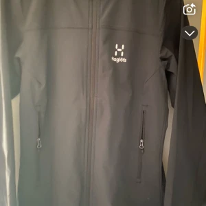 Svart vindjacka från Haglöfs - Svart vindjacka från Haglöfs med stilren design och diskret logga på bröstet. Jackan har huva, två fickor med dragkedja och hel dragkedja framtill. Perfekt för blåsiga dagar och outdoor-aktiviteter. Materialet är syntet och jackan har lång ärm.