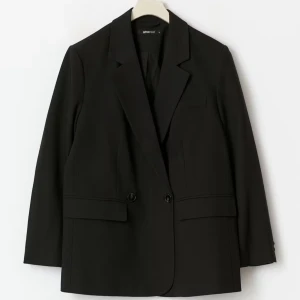 Svart kavaj från Gina Tricot - Stilren svart kavaj från Gina Tricot med klassisk krage och två knappar framtill. Kavajen har raka fickor och en enkel, tidlös design som passar till många olika looks. Perfekt för dig som gillar en clean och snygg stil.