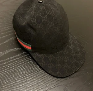 Svart Gucci keps med GG-mönster - Snygg svart keps från Gucci med klassiskt GG-monogram över hela kepsen. Har en grön och röd rand på sidan och justerbar rem baktill i skinn. Materialet är bomull och skinn, och kepsen har en stilren, rundad form som ger en lyxig vibe.