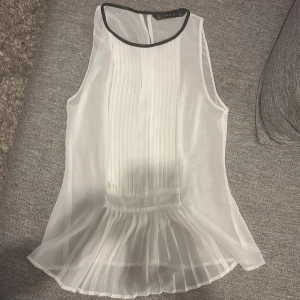 Vit plisserad blus från Zara Trafaluc - Säljer en vit blus från Zara Trafaluc i storlek S. Blusen är ärmlös med svart kant runt halsen, har plisserade detaljer framtill och ett lätt transparent material som ger en luftig känsla. Perfekt för dig som gillar stilrena och eleganta toppar.