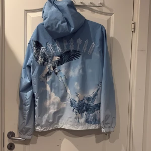 Trapstar blå vindjacka med örntryck - Säljer en blå vindjacka från Trapstar med fett örntryck och moln på både fram och baksidan. Jackan har huva, dragkedja och två fickor med zip. 
