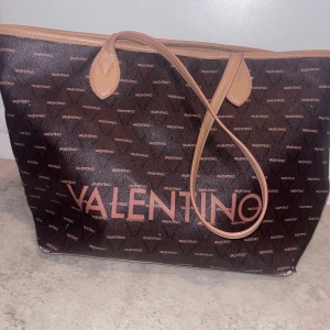 Brun shoppingväska från Valentino - Stor och rymlig shoppingväska från Valentino i mörkbrunt med logomönster över hela väskan. Väskan har ljusbruna detaljer och handtag samt dragkedja upptill. Tillverkad i syntetmaterial med stilren och klassisk design, perfekt för dig som gillar statement-accessoarer.