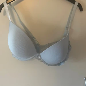 Ljusblå bh med spets från Censored - Säljer en ljusblå bh från Censored med vadderade kupor och fina spetsdetaljer på sidorna. Bh:n har justerbara axelband och en liten rosett framtill. Perfekt för dig som vill ha både komfort och snygg design.