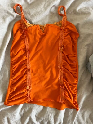 Orange linnetopp med spets och rosetter - Snygg orange linnetopp med smala axelband och dekorativ spets upptill. Toppen har rynkade sidor och små rosetter framtill som ger en söt touch. Perfekt för dig som vill sticka ut med färg och detaljer.