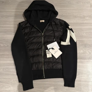 Svart pufferjacka Moncler med huva - Svart pufferjacka från Moncler med huva och snörning. Jackan har quiltad front i syntetmaterial, stickade ärmar med vit broderad detalj och dragkedja framtill. Ribbad nederkant och ärmslut ger en snygg passform. Perfekt för dig som gillar streetstyle. Använd bara 1 månad. Priset kan diskuteras vid snabb affär