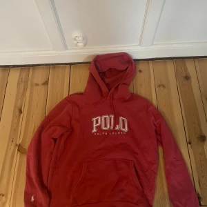 Röd hoodie från Polo Ralph Lauren - Snygg röd hoodie från Polo Ralph Lauren med stor vit logga på bröstet och klassisk huva med snörning. Tröjan har en stor magficka och långa ärmar med broderad logga vid ärmslutet. Perfekt för chill dagar och streetstyle.