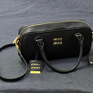 Svart axelväska från Miu Miu i skinn - Snygg svart axelväska från Miu Miu i slätt skinn med guldiga metalldetaljer och dragkedja. Väskan har både handtag och justerbar axelrem samt logga framtill. Perfekt storlek för det viktigaste och stilren design som lyfter varje outfit.