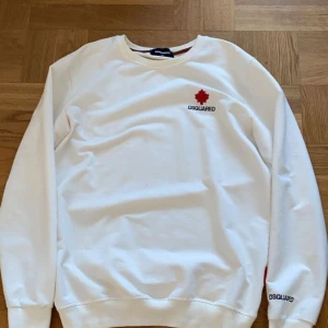 Vit sweatshirt från Dsquared2 - Vit sweatshirt från Dsquared2 med röd lönnlöv-brodyr och svart logotyp på bröstet. Tröjan har rund hals, långa ärmar och ribbade muddar. Materialet är mjuk bomull och det finns en röd Dsquared2-tag vid nederkanten. Perfekt för dig som gillar stilrena designer.