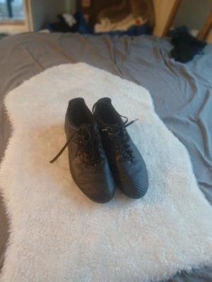 Svarta fotbollsskor med snörning - Säljer ett par svarta fotbollsskor med klassisk rund tå och snörning. Skorna har en stilren look med diskret mönster på ovandelen och platt sula. Perfekta för dig som vill ha en clean och sportig stil på planen.