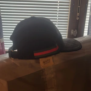 Svart keps från Gucci med mönstrad skärm - Snygg svart keps från Gucci med klassisk röd och grön rand på sidan. Skärmen har ett diskret mönster och kepsen har justerbar rem baktill för perfekt passform. Perfekt för dig som vill ha en stilren och lyxig accessoar.