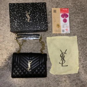 Svart quiltad axelväska från YSL - Snygg svart axelväska från YSL i quiltat skinn med guldiga kedjedetaljer och ikonisk YSL-logga framtill. Klassisk rektangulär form och elegant design som verkligen sticker ut. Levereras med originalbox och dustbag.