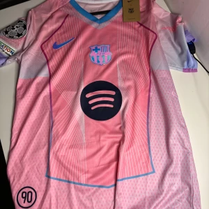 Barça x Nike rosa fotbollströja - Snygg FC Barcelona fotbollströja från Nike i rosa och ljusblå toner. Tröjan har korta ärmar, V-ringning med blå kant, tryckt klubbmärke, Champions League-märke och stor svart Spotify-logga på bröstet. Tillverkad i ventilerande polyester för max komfort.