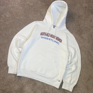 Vit hoodie Russell Athletic wrestling - Säljer en vit vintage hoodie från Russell Athletic med trycket 'Northfield Mount Hermon Wrestling' i rött och blått på bröstet. Tröjan har huva med snörning, stor magficka och långa ärmar. Perfekt för dig som gillar sportig och avslappnad stil.