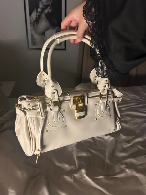 Beige paddington handväska  - Snygg beige paddington handväska i skinn med guldfärgade metalldetaljer, lås och nitar. Väskan har dubbla handtag, dekorativa sömmar och spännen samt dragkedja upptill. Inte äkta!! 🩷