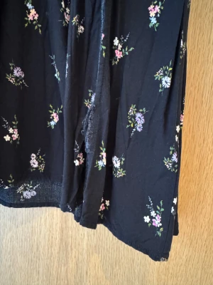 Svart blommig långklänning H&M - Svart långklänning från H&M DIVIDED i storlek XS med små blommor i rosa och vitt. Klänningen har v-ringning, smock upptill, långa ärmar med resår och en snygg slits framtill. Materialet är mjukt och lätt polyester, perfekt för dig som gillar romantisk stil. Blommig långärmad klänning med slits och volangkant. Tråd har släppt lite i slitsen (se bild).