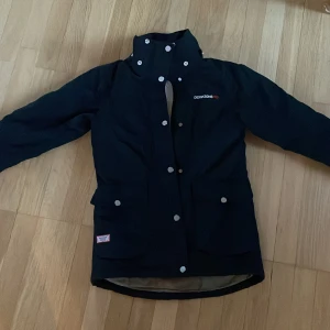 Svart teddyjacka från Didriksons - Svart teddyjacka från Didriksons med hög krage och stora fickor framtill. Jackan har vita knappar och ett mjukt, vitt teddyfoder på insidan som gör den extra skön. Märkeslogga broderad på bröstet och patch på fickan. Perfekt för kyliga dagar.