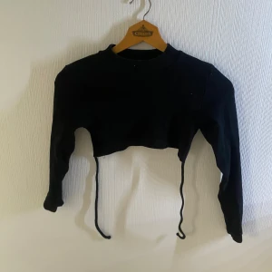 Svart croppad långärmad topp - Svart croppad topp med långa ärmar och snörning framtill. Enkel och stilren design som passar perfekt till höga jeans eller kjol. Mjuk och stretchig känsla för en bekväm passform.