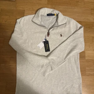 Ljusgrå half zip tröja Polo Ralph Lauren - Snygg ljusgrå långärmad tröja från Polo Ralph Lauren med broderad logga i brunt på bröstet. Tröjan har en stilren half zip-dragkedja med läderdetalj och klassisk krage. Perfekt för dig som gillar enkel och clean stil.
