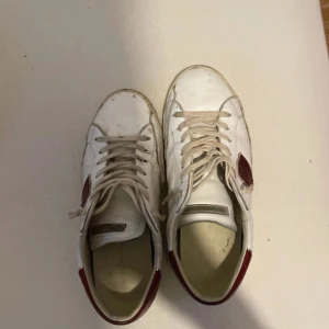 Philippe Model vita sneakers med röda detaljer - Snygga vita sneakers från Philippe Model med röda detaljer på häl och sida. Klassisk låg modell med snörning och rund tå. Skorna är i skinn och har en platt sula. Perfekta för dig som gillar stilrena men ändå unika sneakers.