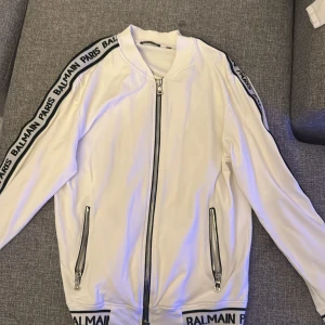 Vit kofta från Balmain Paris - Snygg vit tröja från Balmain Paris med svarta logoband längs ärmar och nederkant. Jackan har ribbad krage, två fickor med dragkedja och en kraftig dragkedja framtill. Materialet är mjukt och känns som polyester. Perfekt för dig som gillar streetwear med lyxig vibe.