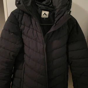 Svart pufferjacka från McKinley - Svart pufferjacka från McKinley i storlek 36. Jackan har quiltad design, huva och dragkedja framtill. Perfekt för kalla dagar med sin tjocka och fluffiga look. Passformen är normal och jackan har långa ärmar. Snygg och enkel stil som funkar till det mesta. (ej använd)