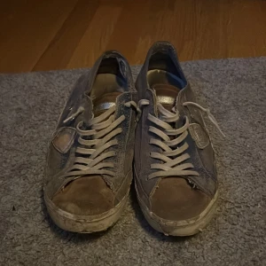 Philippe Model grå sneakers i mocka - Säljer ett par Philippe Model sneakers i grå mocka med vita snören och detaljer i silver. Skorna har en klassisk låg profil, rund tå och platt sula. Sidan pryds av en silverfärgad sköld och sulan har märkets logga undertill. Perfekta för en avslappnad streetstil.