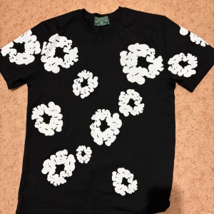 Denim tests t- shirt  - Cool svart t-shirt från Denim Teats med stora vita blommiga tryck över hela tröjan. Klassisk passform och rund halsringning. Perfekt för dig som vill sticka ut med en unik och trendig look.