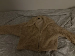Beige teddyjacka med dragkedja - Säljer en beige teddyjacka med mjukt fluffigt material och krage. Jackan har en dragkedja framtill och är croppad i modellen, perfekt för dig som gillar en chill och cozy stil. Långärmad och passar till lager-på-lager under kyliga dagar.