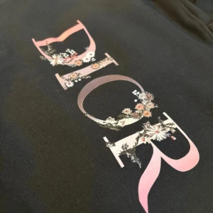 Svart Dior hoodie med blommigt tryck - LÄS HELA! Svart hoodie från Dior i storlek M med stort blommigt Dior-tryck i rosa och vitt på bröstet. Tröjan har huva med snörning och en klassisk magficka. Tillverkad i 100% bomull för skön och mjuk känsla. Perfekt för dig som vill ha en lyxig streetwear-look. PRIS DISKUTERAS, DÄRAV PRISET