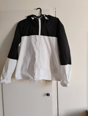 Svartvit vindjacka från FB Sister - Svartvit vindjacka från FB Sister med huva och snörning. Jackan har dragkedja framtill, två sidofickor med dragkedja och elastisk nederkant. Materialet är syntet och jackan har en loose passform. Perfekt för dig som gillar sportig stil.