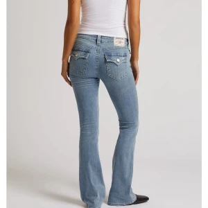 True Religion ljusblå bootcut jeans - Snygga ljusblå jeans från True Religion i modellen Becca Mid Rise Bootcut Flap. Jeansen har medelhög midja, klassisk bootcut och dekorativa ficklock med knappar bak. Perfekt passform och coola detaljer som ger en trendig look. Säljer dem för de aldrig kommer till användning 