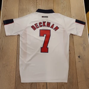 England Beckham 7 fotbollströja Umbro - Klassisk England fotbollströja från Umbro med Beckham och nummer 7 på ryggen. Tröjan är vit med marinblå krage, röda och blå detaljer på sidor och ärmar samt broderat landslagsmärke på bröstet. Tillverkad i lätt och ventilerande material.
