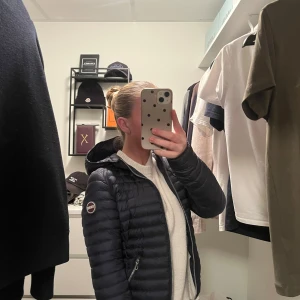 Mörkblå dunjacka från Colmar - Säljer en snygg mörkblå dunjacka från Colmar med huva och quiltad design. Jackan har dragkedja framtill och två praktiska fickor med dragkedja. Den är i storlek 40 men den passar mig perfekt som alltid har xs på mig, så skulle säga att denna modell i storlek 40 motsvarar xs