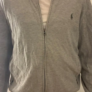 Grå zipjacka från Polo Ralph Lauren - Snygg grå zipjacka från Polo Ralph Lauren med klassisk logga på bröstet. Jackan har två fickor med dragkedja och är tillverkad i mjuk bomull. Perfekt för chill dagar och enkel att matcha med jeans eller joggers.