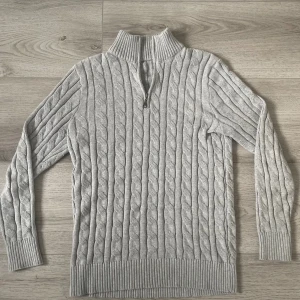 Grå kabelstickad tröja med half zip - Säljer en grå kabelstickad tröja med ribbad krage och half zip-dragkedja. Tröjan är tillverkad i 100% bomull och har långa ärmar samt breda ribbade muddar vid ärmslut och nederkant. Perfekt för dig som gillar klassisk stil med en modern twist. Det är storlek M. Skriv för fler frågor 