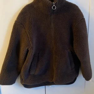 Brun teddyjacka från Etirel - Mysig brun teddyjacka från Etirel i storlek S. Jackan har en hög krage, dragkedja med rund metallring och två sidofickor. Materialet är fluffigt och ger en chill oversized vibe, perfekt för kalla dagar. Enkel design utan mönster och med långa ärmar.