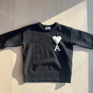 Svart stickad tröja från AMI Paris - Svart stickad tröja från AMI Paris med vit hjärtformad detalj och bokstaven A på bröstet. Rund halsringning och ribbade muddar vid ärmslut och nederkant. Perfekt för dig som gillar stilrena och coola plagg med en twist.