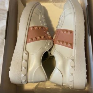 Valentino Rockstud sneakers vit/rosa - Snygga Valentino Rockstud sneakers i vitt skinn med ljusrosa band och ikoniska nitar längs sidorna och sulan. Rund tå, platt sula och snörning framtill. Perfekt för dig som gillar lyxiga detaljer och modern design. Kom privat för flera bilder på skorna då jag har använt dom. 