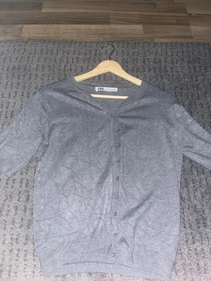 Grå kofta från Zara med knappar - Grå kofta från Zara i storlek M. Den har en klassisk rund halsringning och knappar hela vägen framtill. Materialet är mjukt och tunt, perfekt för lager-på-lager. Ärmarna är lite kortare än vanliga långärmade och koftan har ribbade muddar vid nederkant och ärmslut.