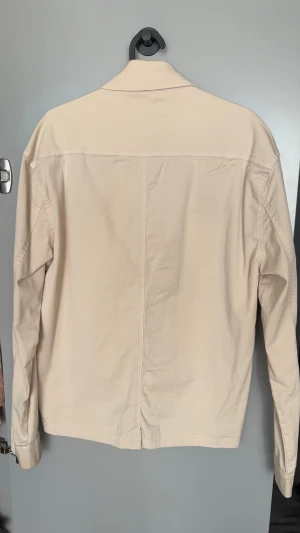 Beige overshirt från Studio Total - Snygg beige overshirt från Studio Total med klassisk krage, stora frontfickor och knäppning med bruna knappar. Jackan är tillverkad i mjuk bomull och har en clean, minimalistisk look som passar till många. 