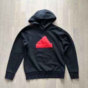 Svart hoodie från Adidas med röd logga - Säljer en svart hoodie från Adidas med stor röd logga på bröstet. Tröjan har huva, långa ärmar och breda muddar vid ärmslut och nederkant. Tillverkad i mjuk bomullsmix som är skön att ha på sig. Perfekt för dig som gillar sportig och clean stil. 