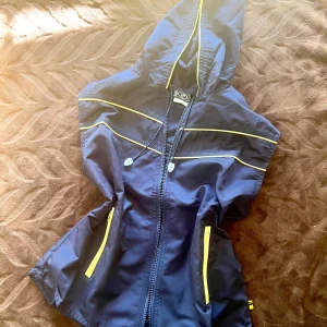 Blå vindjacka med gula detaljer - Snygg marinblå vindjacka från ICD med huva och gula detaljer längs dragkedjor och sömmar. Jackan har två fickor med gul dragkedja och justerbar huva med snörning. Perfekt för blåsiga dagar och en sportig look. Funkar även för träning 