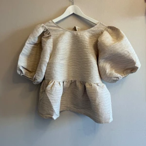 Beige blus med puffärm Carin Wester - Supersnygg beige blus från Carin Wester med voluminösa puffärmar och rynkad midja. Blusen har en snygg struktur i tyget och en knytdetalj i ryggen som detalj. Perfekt för dig som gillar statement-ärmar och en feminin look. Helt ny! Rökfritt hem.