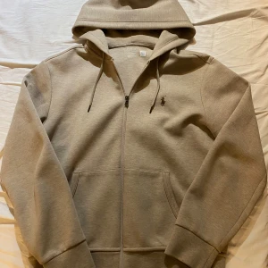 Beige hoodie från Polo Ralph Lauren - Snygg beige hoodie från Polo Ralph Lauren med huva, snörning och dragkedja framtill. Klassisk logga broderad på bröstet och två stora fickor. Perfekt för chill dagar och enkel att matcha med jeans eller joggers.