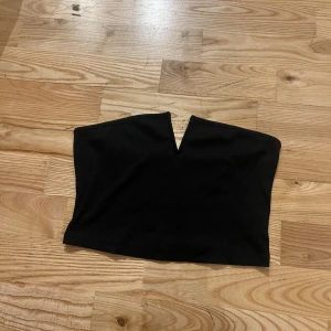 Svart bandeau topp - Säljer en svart bandeau tubtopp med snygg V-ringad detalj framtill. Toppen är croppad i ribbat material. Använd enstaka gång.