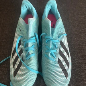 Adidas fotbollsskor - Säljer ett par Adidas X 19.3 fotbollsskor i ljusblått med svarta ränder. Skorna har snörning och en platt, rund tå. Yttermaterialet är syntet och sulan har vita dobbar för bra grepp på planen. 